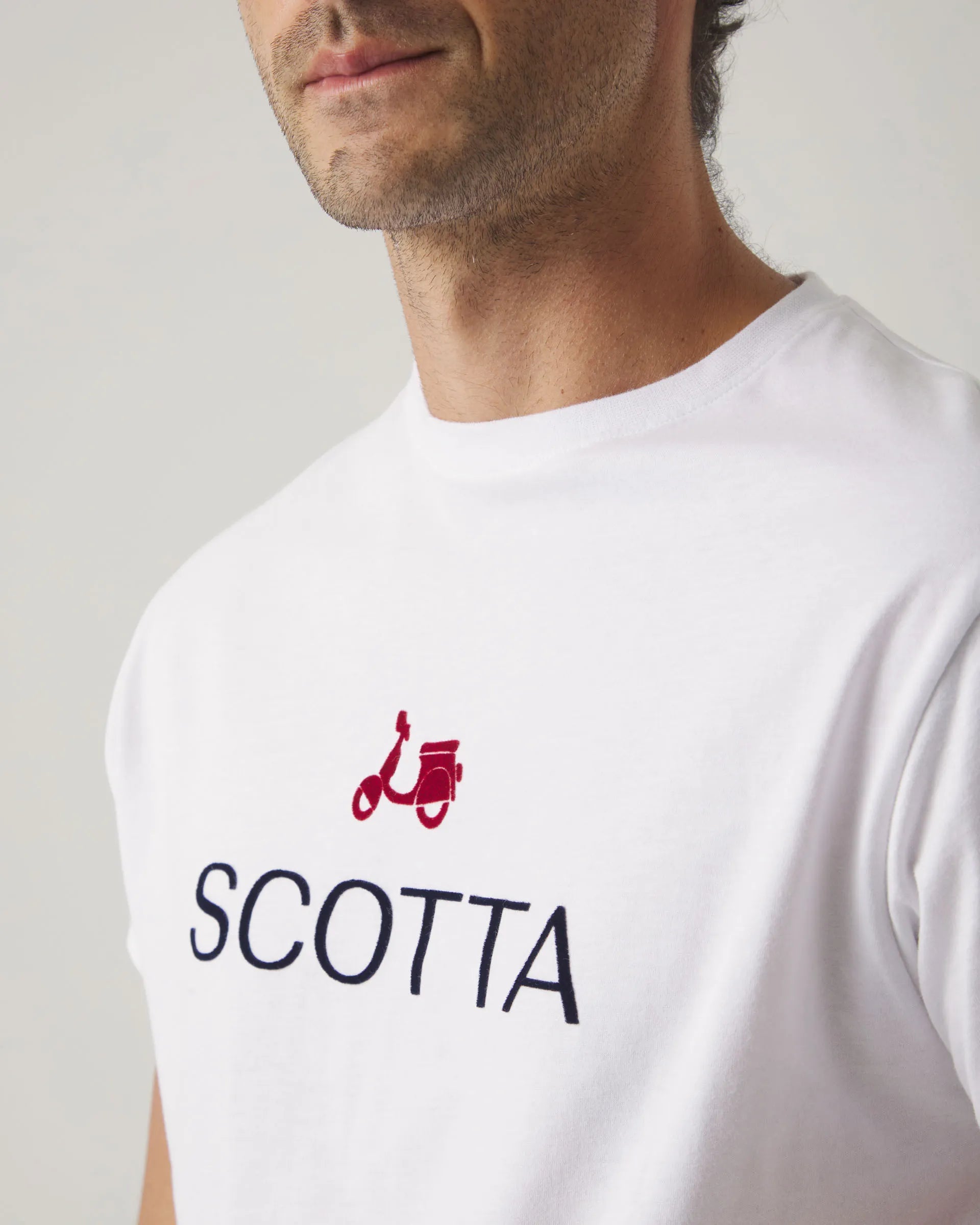Camiseta blanca con cuello redondo y manga corta, con letras y moto Scotta en el torso.